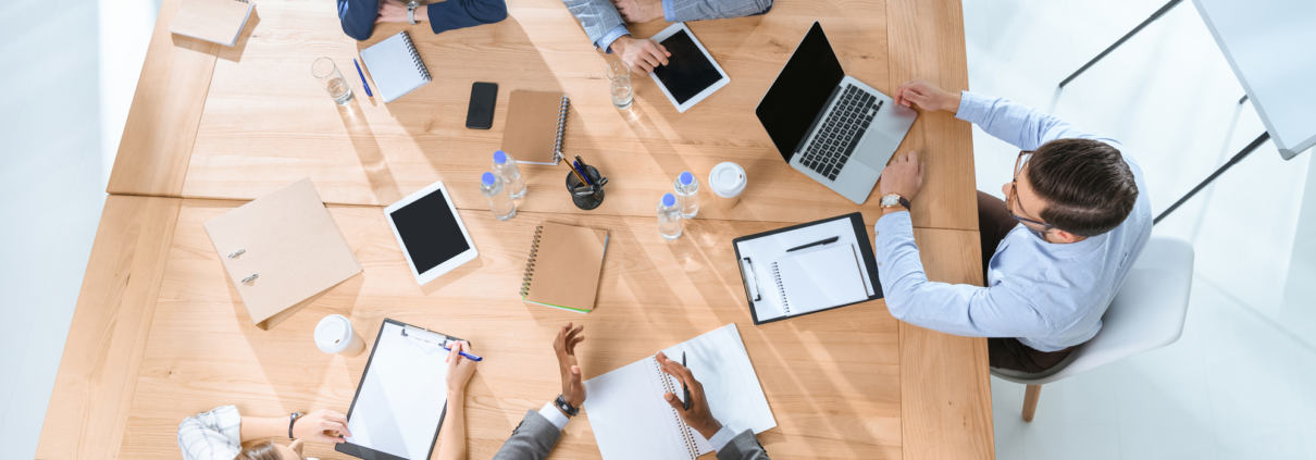 Eine Gruppe von fünf Personen sitzt an einem Tisch und arbeitet zusammen mit Notizbüchern, Tablets und Laptops.