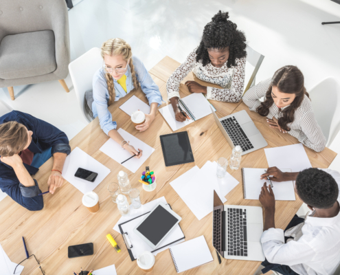 Eine Gruppe von Menschen sitzt an einem Tisch, arbeitet zusammen und macht Notizen mit Laptops, Tablets und Schreibmaterial.