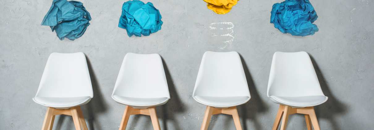 Vier weiße Stühle vor einer Wand mit bunten Papierknäueln, die eine Glühbirne symbolisieren.