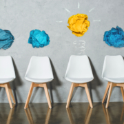 Vier weiße Stühle vor einer Wand mit bunten Papierknäueln, die eine Glühbirne symbolisieren.