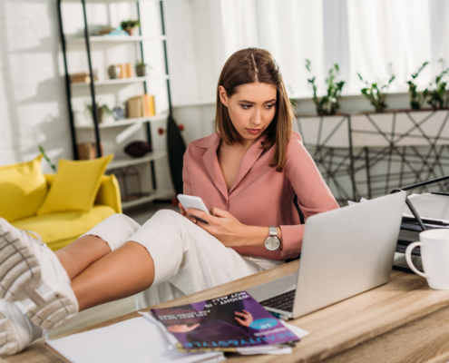 Eine Frau sitzt entspannt in einem modernen Büro, hat die Füße auf dem Tisch, hält ein Smartphone und schaut auf einen Laptop.