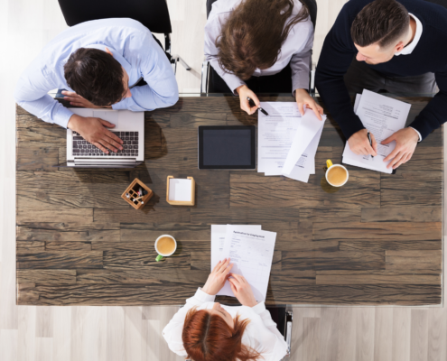 Vier Personen arbeiten an einem Tisch mit Laptops und Dokumenten. Kaffeetassen und Schreibutensilien sind verteilt.