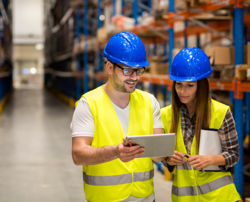 Zwei Personen in Schutzhelmen und Warnwesten stehen in einem Lager und schauen gemeinsam auf ein Tablet.