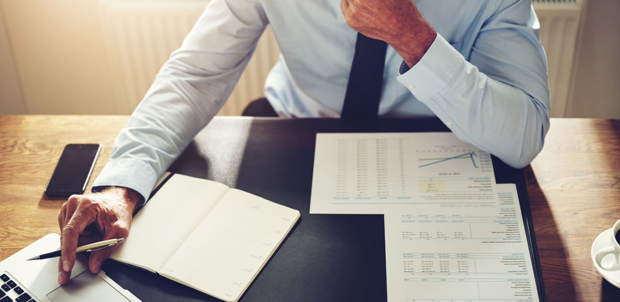 Eine Person in einem Hemd sitzt an einem Schreibtisch und arbeitet mit Dokumenten, einem Notizbuch und einem Laptop.
