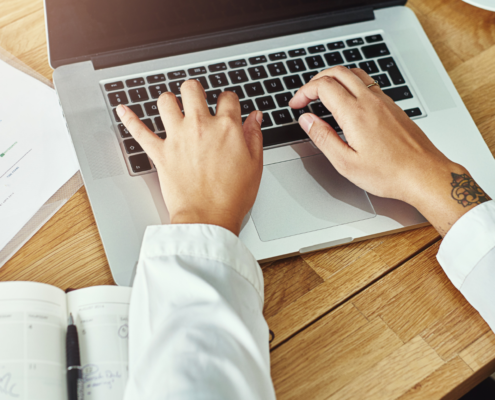Person tippt auf einem Laptop, mit einem Kalender und Diagrammen auf dem Schreibtisch daneben.
