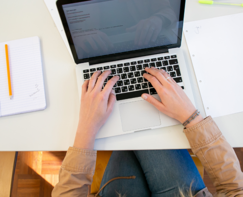 Eine Person arbeitet an einem Laptop mit Notizbüchern und Papieren auf dem Schreibtisch.