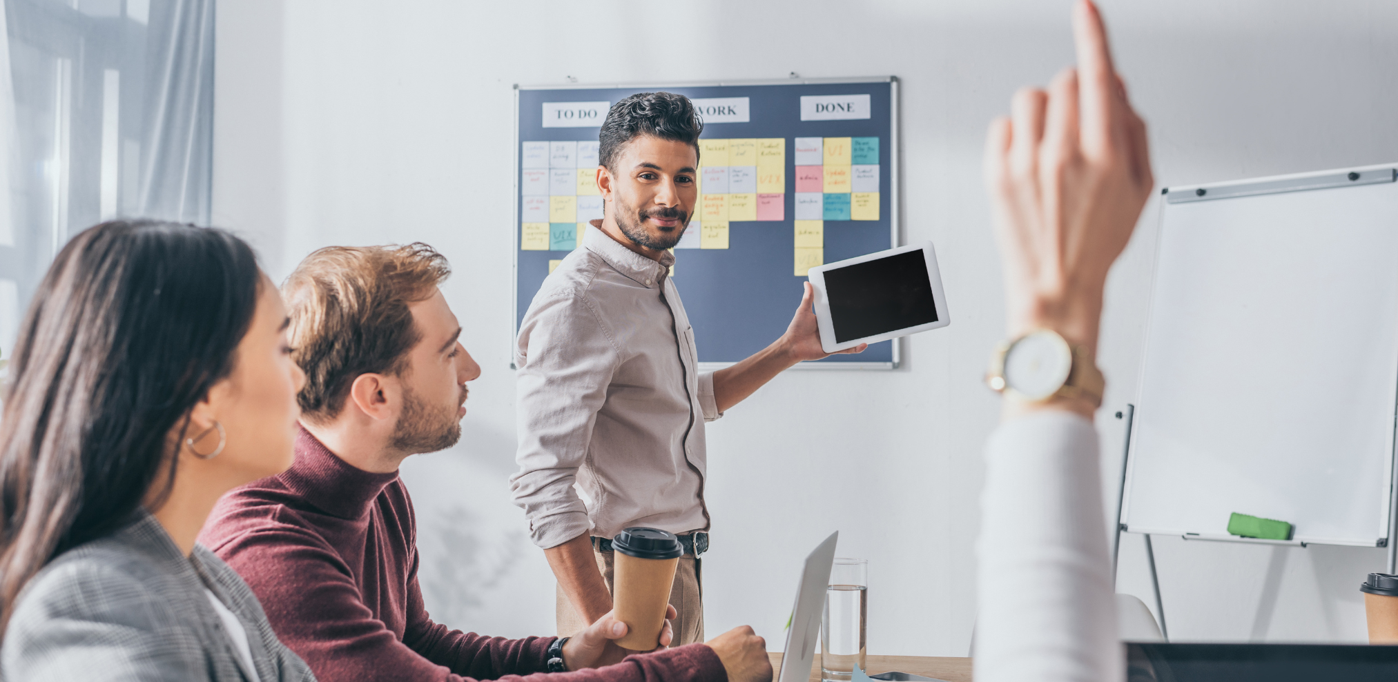Ein Mann hält eine Präsentation und zeigt dabei auf ein Tablet, während er vor einer Gruppe von Kollegen steht, die ihm aufmerksam zuhören. Im Hintergrund ist ein Kanban-Board mit Post-its zu sehen.