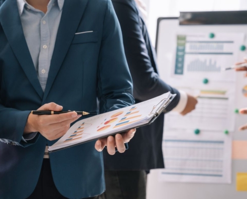 Geschäftspersonen besprechen Diagramme auf einem Clipboard, während andere auf eine Tafel mit Diagrammen und Notizen im Hintergrund zeigen.
