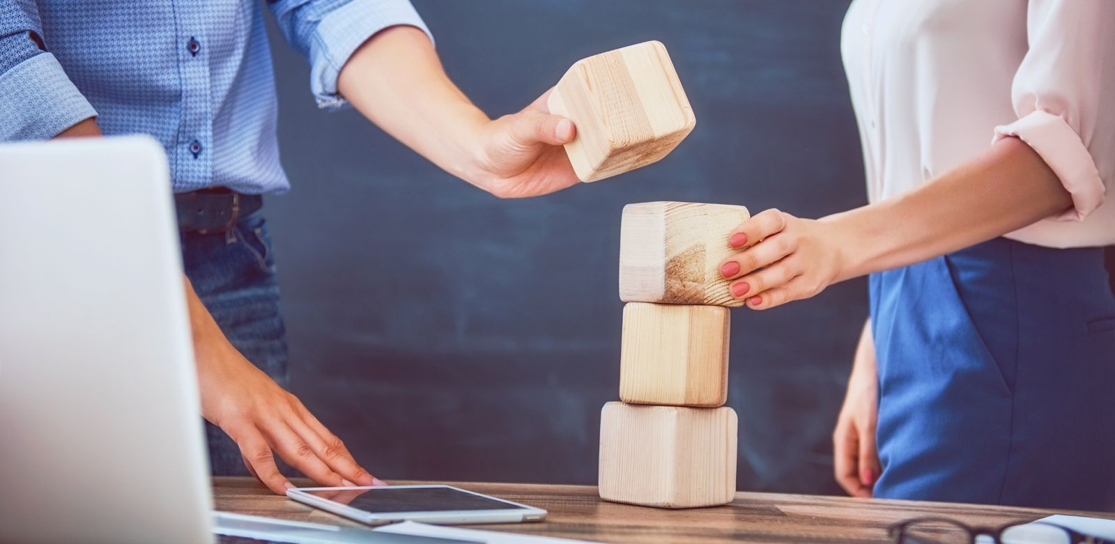 Zwei Personen stapeln gemeinsam Holzblöcke auf einem Tisch, auf dem ein Tablet liegt.