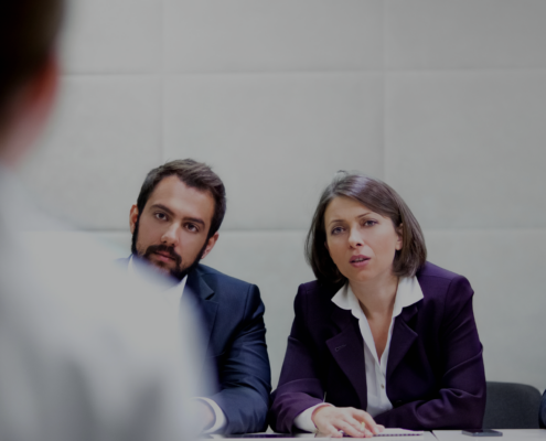 Drei Personen in Geschäftskleidung sitzen an einem Tisch und hören einer vierten Person zu, die außerhalb des Bildes steht und spricht. Die Zuhörer wirken aufmerksam und engagiert.