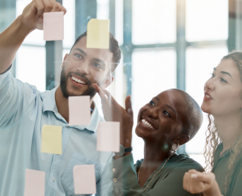 Drei Personen arbeiten gemeinsam und befestigen Haftnotizen an einer Glaswand. Sie lachen und diskutieren dabei.