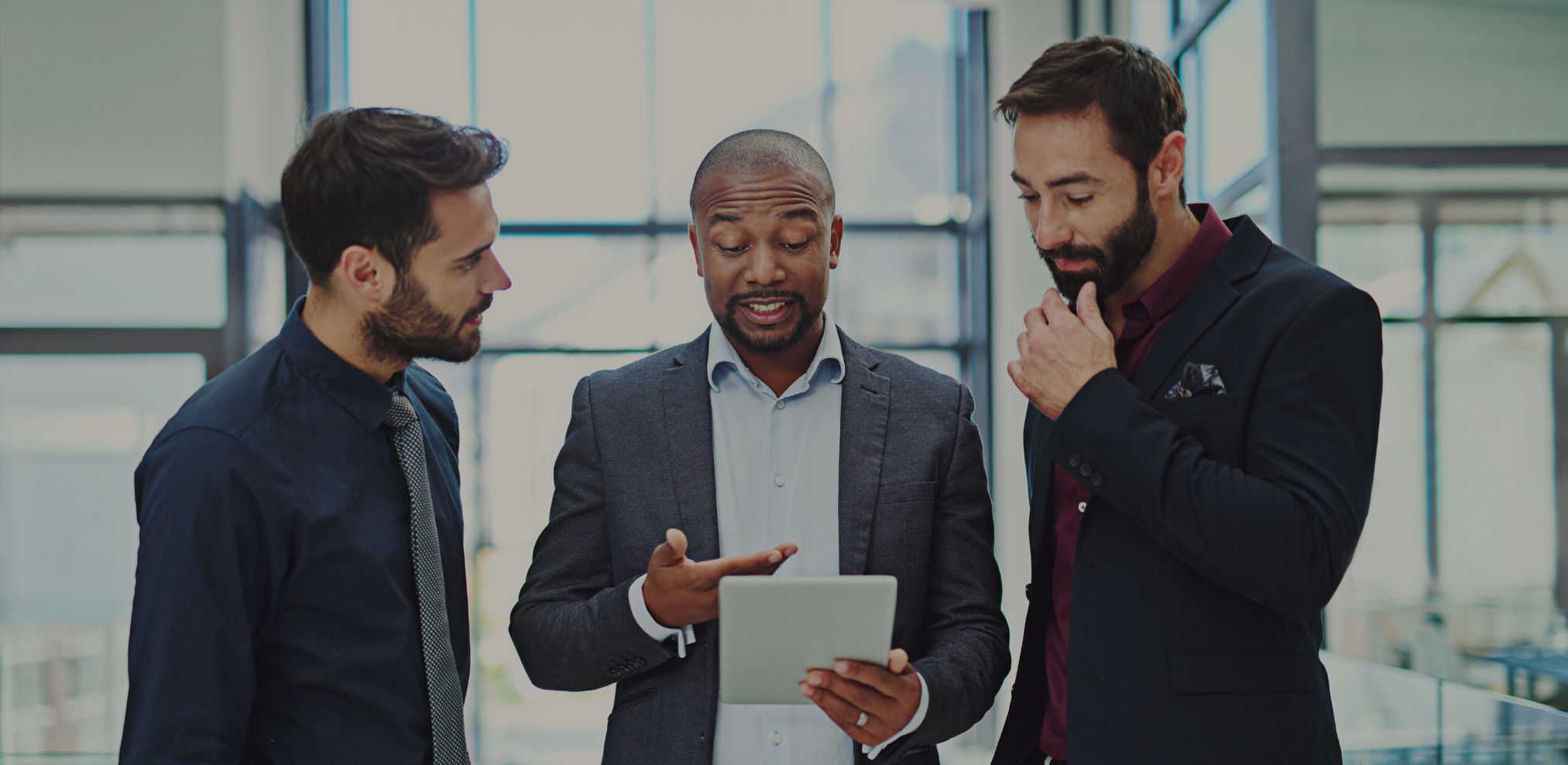 Drei Männer in Geschäftskleidung stehen zusammen und diskutieren. Einer der Männer hält ein Tablet und spricht, während die anderen beiden zuhören.