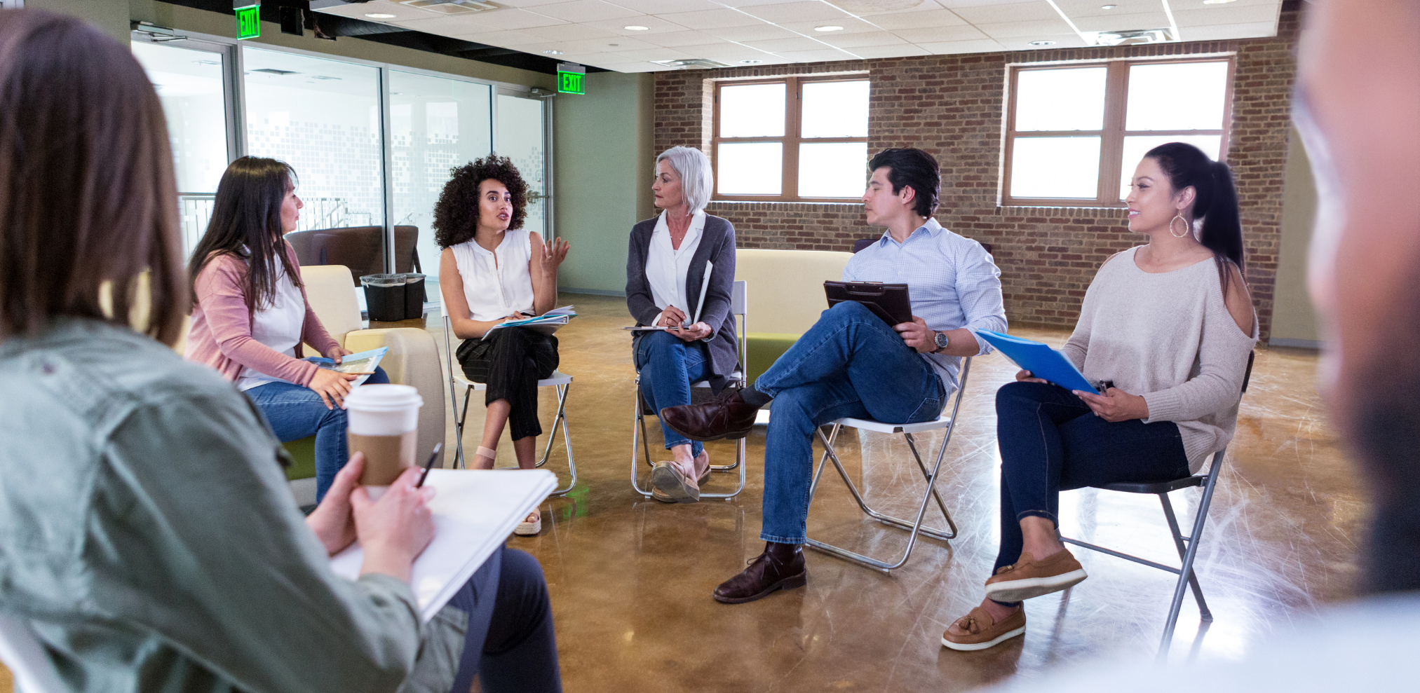 Eine Gruppe von Menschen sitzt in einem Kreis und diskutiert. Eine Person spricht gestikulierend, während die anderen aufmerksam zuhören und Notizen machen.