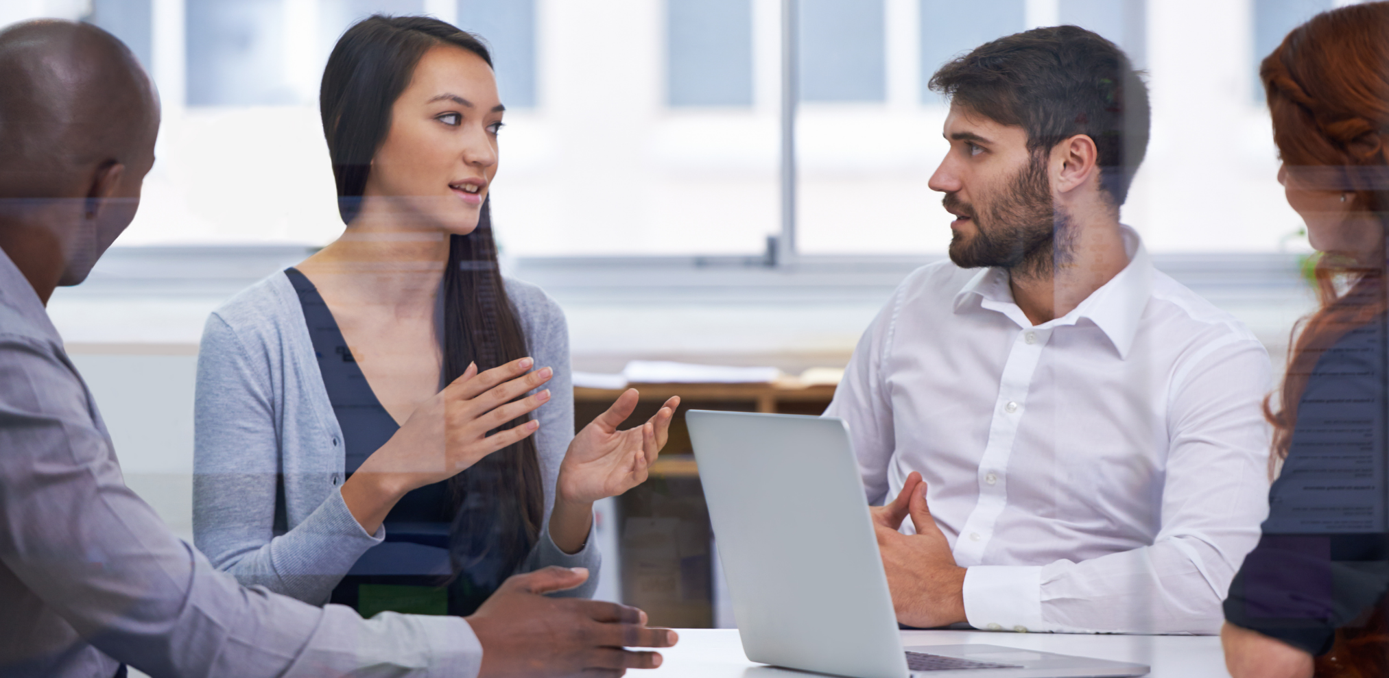 Vier Personen sitzen an einem Tisch und diskutieren. Eine Frau spricht und gestikuliert, während die anderen aufmerksam zuhören. Auf dem Tisch steht ein Laptop.