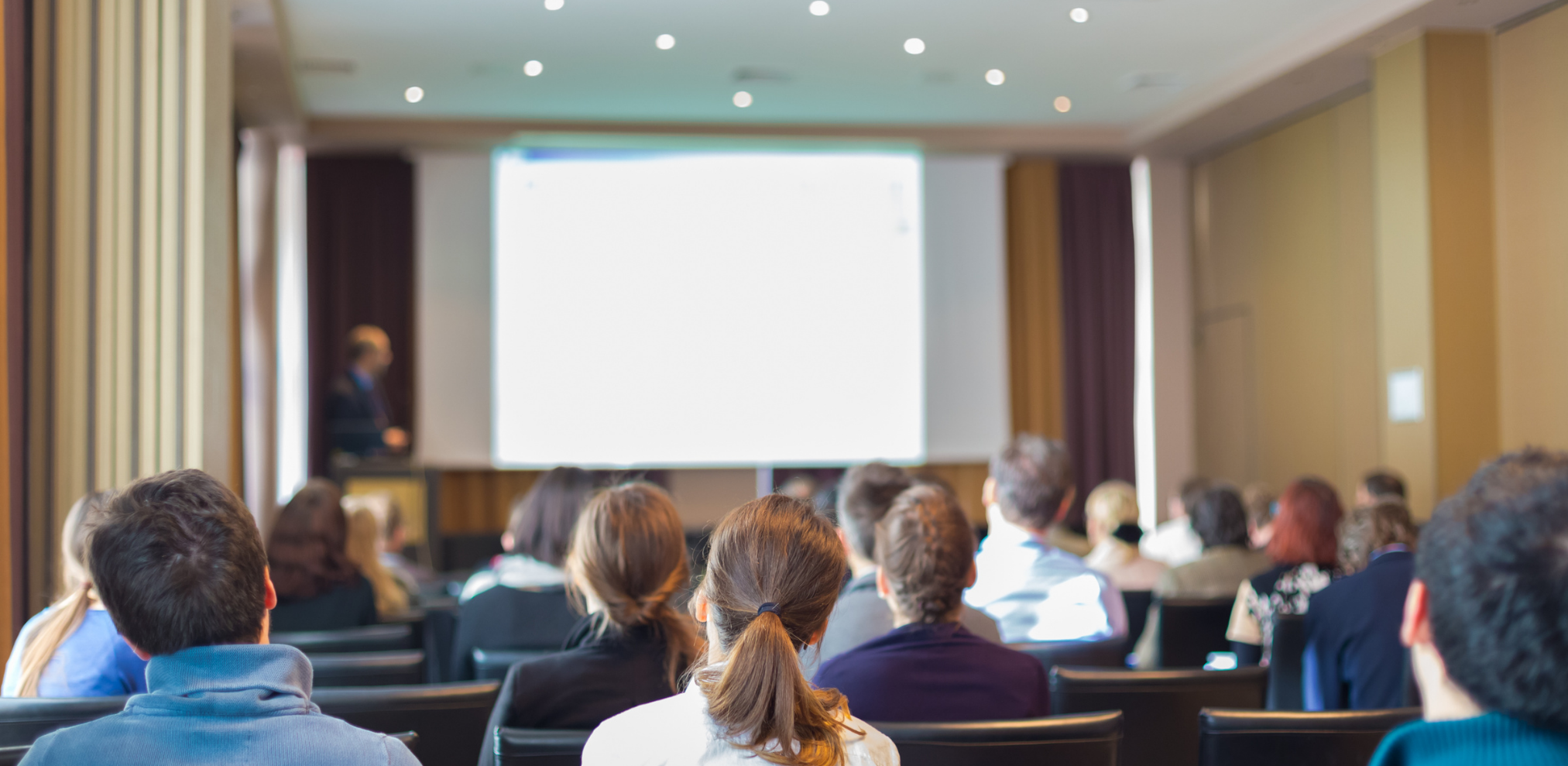 Ein Vortragssaal mit Menschen, die einem Vortrag zuhören. Im Vordergrund sind die Rücken der Zuschauer zu sehen, während vorne ein unscharfer Präsentator vor einer Leinwand steht.