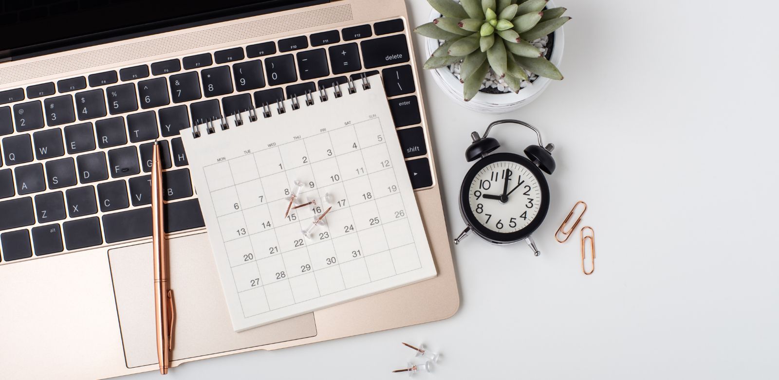 Ein Schreibtisch mit einem Laptop, einem Kalender, einem Stift, einer Uhr, Büroklammern und einer kleinen Pflanze.