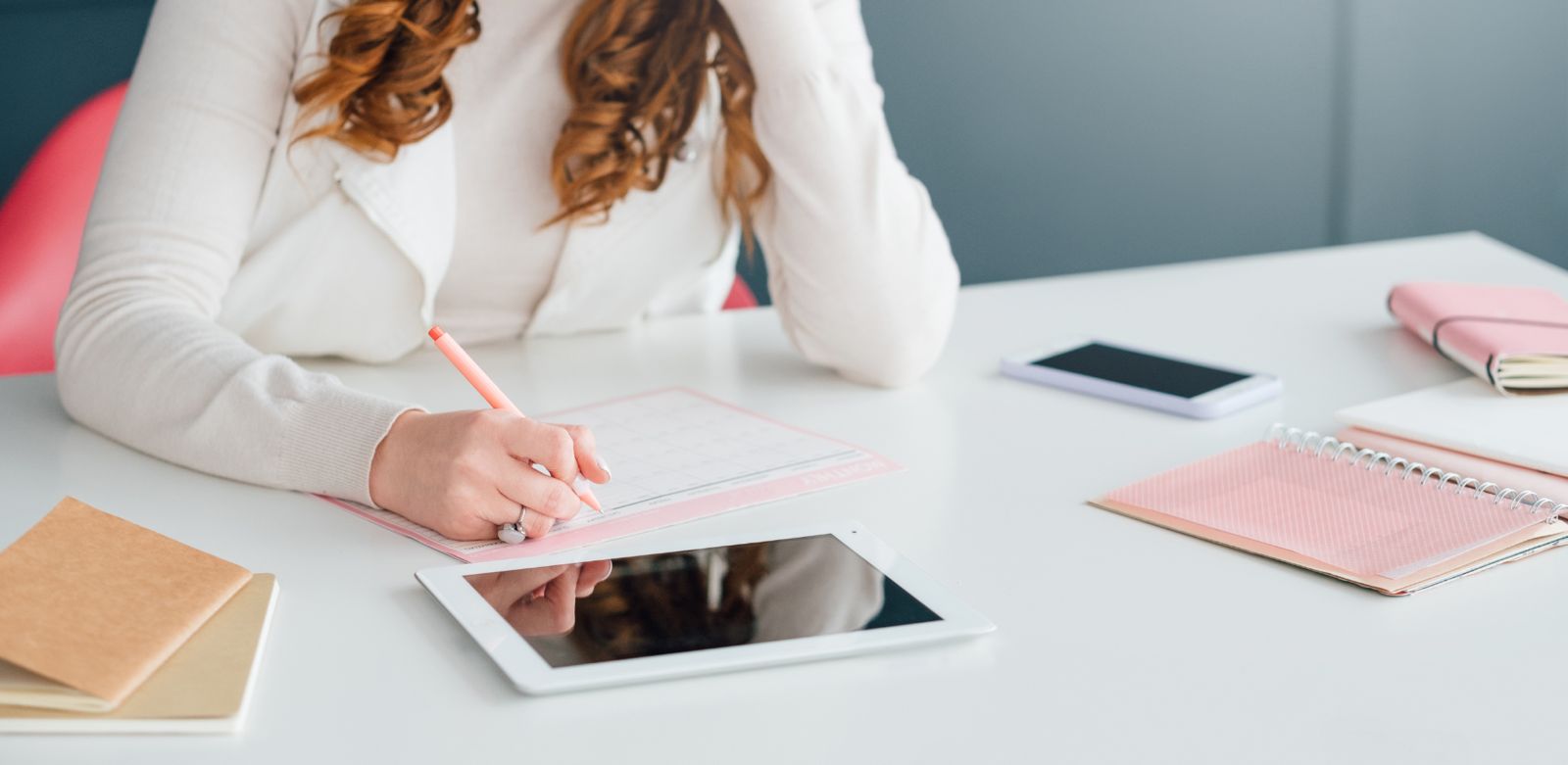 Eine Person sitzt an einem Schreibtisch und schreibt in einen Planer, umgeben von einem Tablet, einem Smartphone und Notizbüchern.