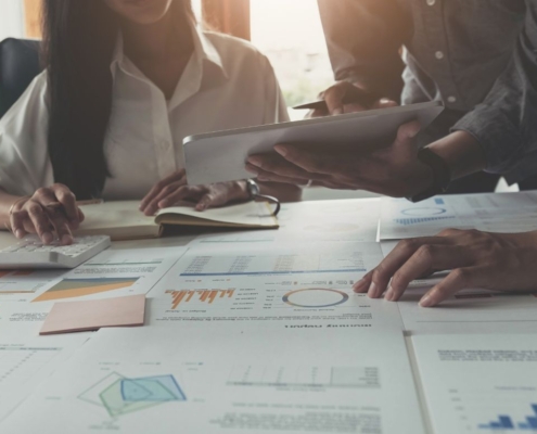 Mehrere Personen arbeiten an einem Tisch, der mit Finanzberichten und Diagrammen bedeckt ist, während sie mit einem Tablet und einem Taschenrechner arbeiten.