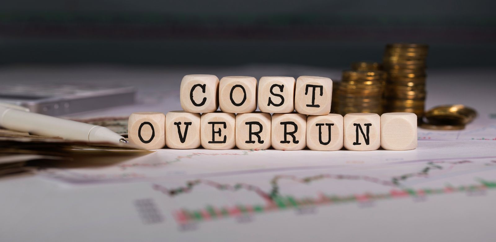 Holzwürfel mit der Aufschrift "COST OVERRUN" auf einem Tisch mit Finanzdokumenten und Münzstapeln im Hintergrund.