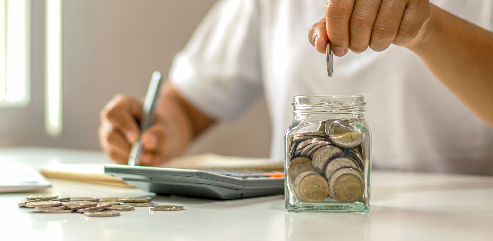 Eine Person zählt Münzen und gibt sie in ein Glas, während sie neben einem Taschenrechner sitzt und Notizen macht.