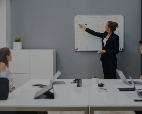 Eine Person steht vor einem Whiteboard und erklärt mehreren sitzenden Kollegen etwas in einem Konferenzraum.