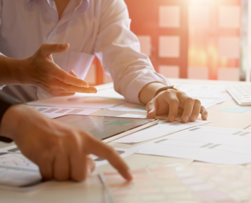 Zwei Personen arbeiten an einem Schreibtisch und diskutieren über Dokumente und Diagramme, die auf dem Tisch verteilt sind. Eine Person zeigt auf ein Papier, während die andere ihre Hand über ein Tablet hält.