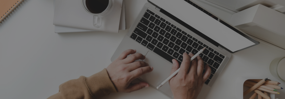 Das Bild zeigt eine Person, die an einem Laptop arbeitet. Auf dem Schreibtisch befinden sich eine Tasse Kaffee, ein Notizbuch, Bleistifte und ein Stifthalter. Die Person hält einen Stift in der rechten Hand und tippt mit der linken Hand auf der Tastatur.