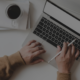 Das Bild zeigt eine Person, die an einem Laptop arbeitet. Auf dem Schreibtisch befinden sich eine Tasse Kaffee, ein Notizbuch, Bleistifte und ein Stifthalter. Die Person hält einen Stift in der rechten Hand und tippt mit der linken Hand auf der Tastatur.