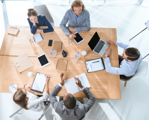 Eine Gruppe von fünf Personen sitzt an einem Tisch und arbeitet zusammen mit Notizbüchern, Tablets und Laptops.