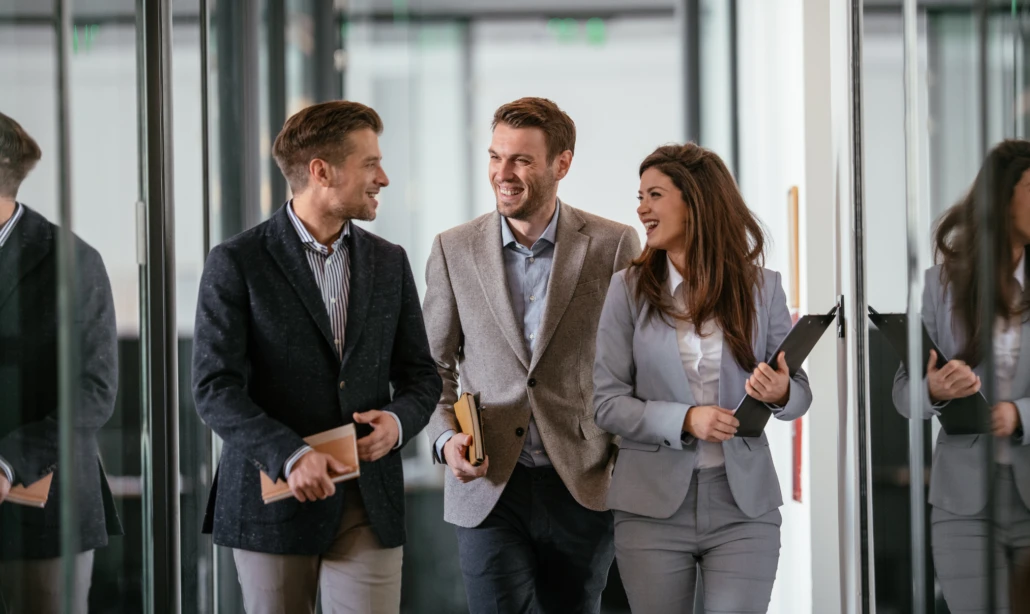 Drei Geschäftsleute gehen lachend durch einen Büroflur, während sie miteinander sprechen.