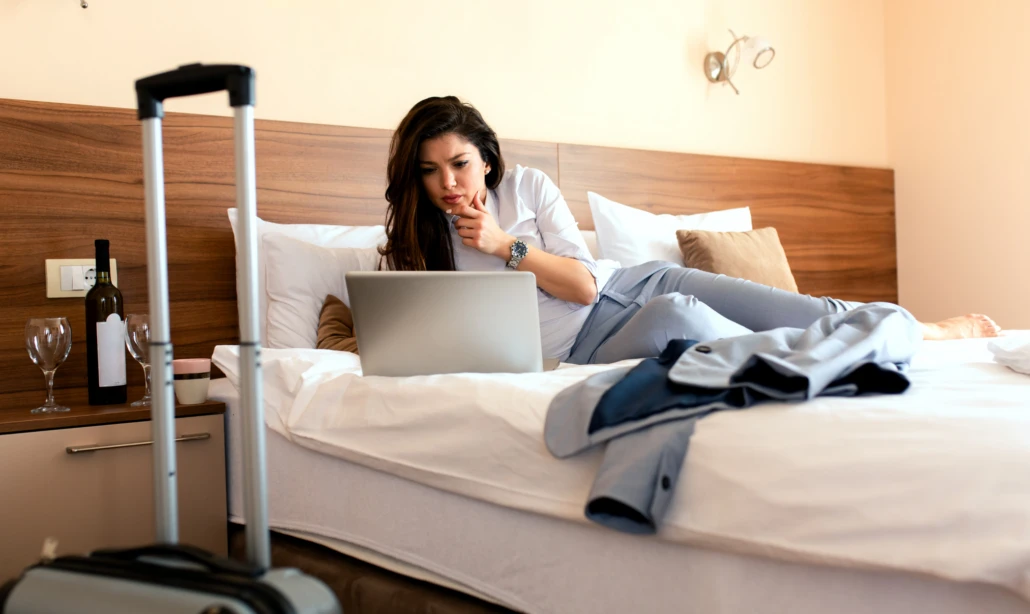 Frau arbeitet in einem Hotelzimmer am Laptop, während ein Rollkoffer und eine Weinflasche im Vordergrund stehen.