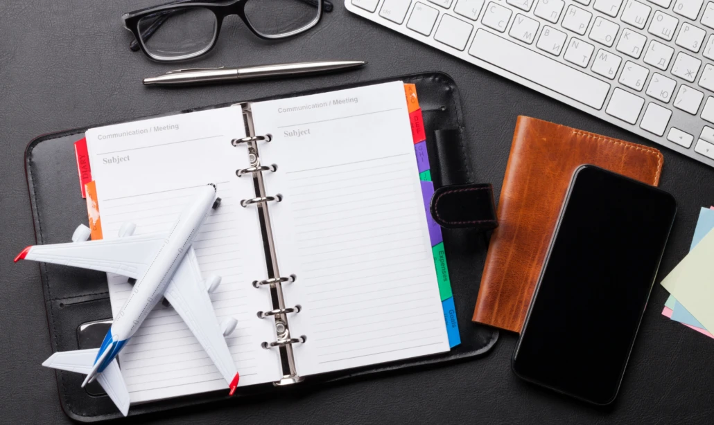 Schreibtisch mit einem Notizbuch, Modellflugzeug, Brille, Stift, Smartphone, Geldbörse und einer Tastatur.