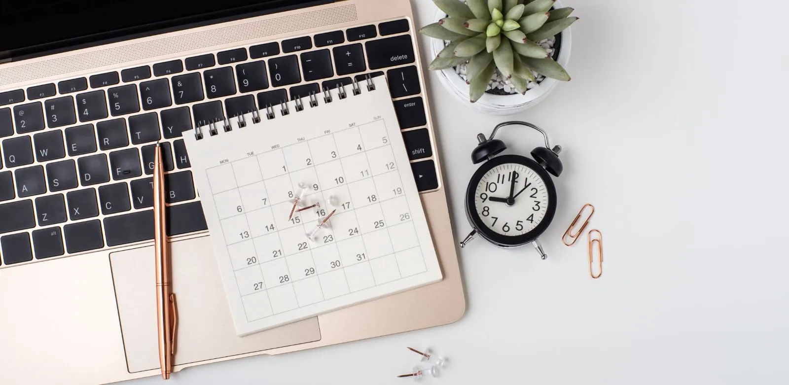 Ein Schreibtisch mit einem Laptop, einem Kalender, einem Stift, einer Uhr, Büroklammern und einer kleinen Pflanze.