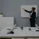 Eine Person steht vor einem Whiteboard und erklärt mehreren sitzenden Kollegen etwas in einem Konferenzraum.